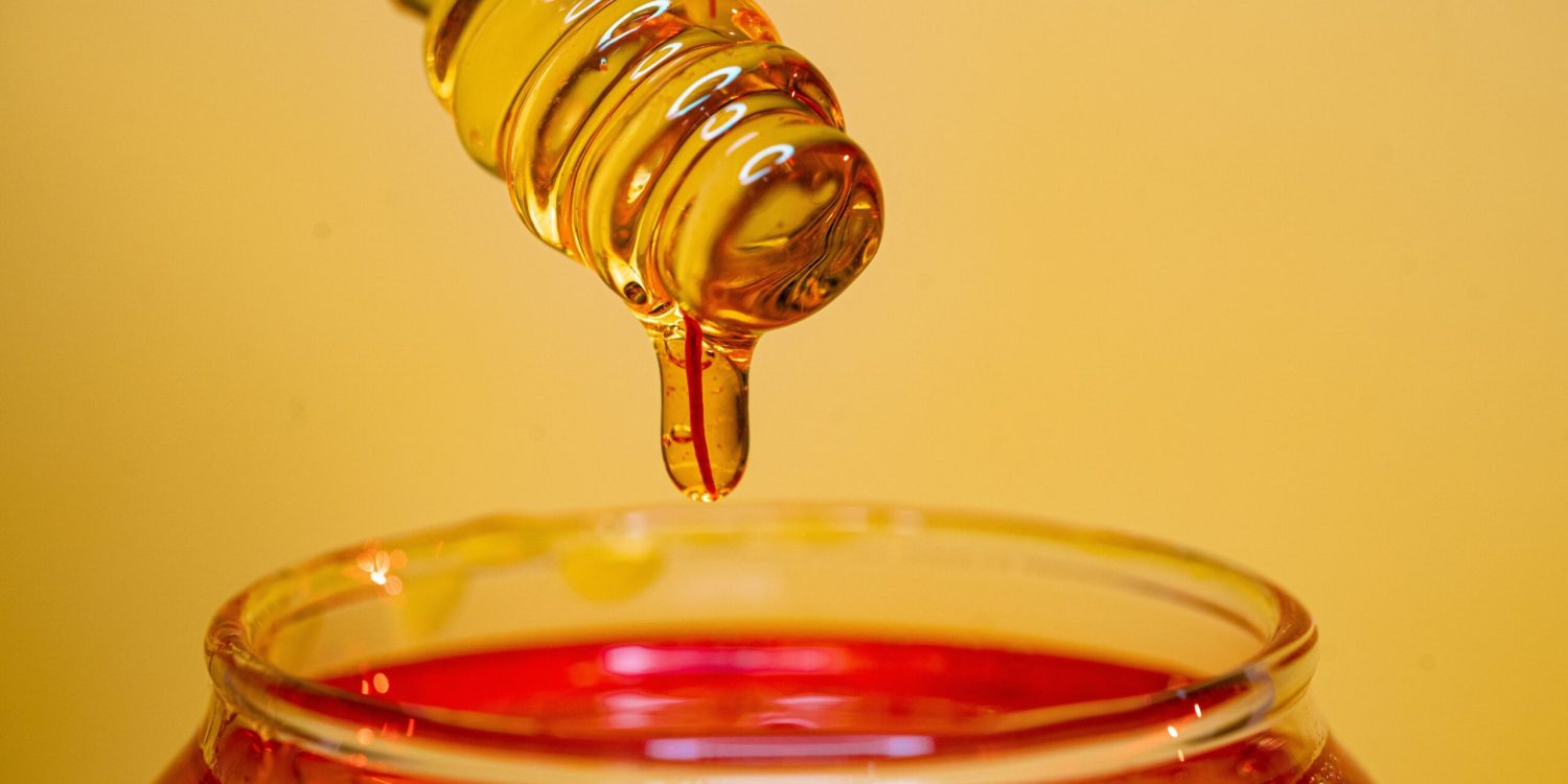 A spoon drips golden honey into a clear glass, creating a sweet and sticky cascade.