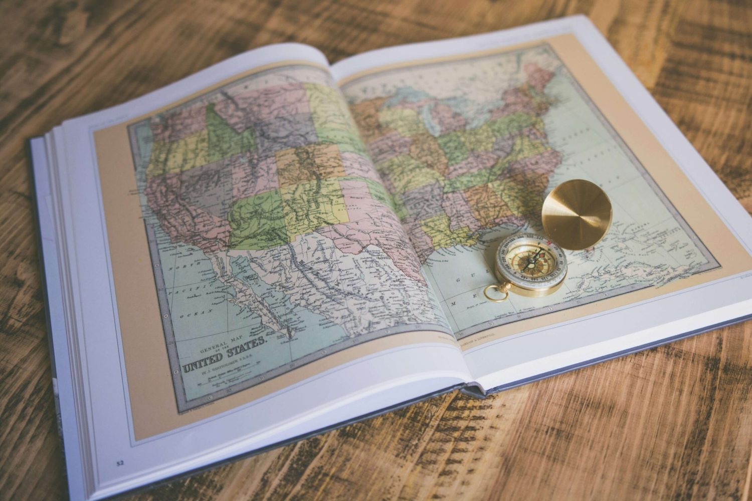 Open book displaying a map of the United States, with a gold compass resting on the right page. The book is on a wooden surface.